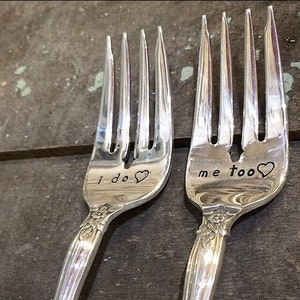 Puede incluir: Dos tenedores de plata con las palabras "Yo sí" y "Yo también" grabadas, cada uno con un pequeño símbolo de corazón.