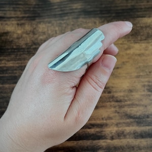 May include: A long, silver-toned metal ring with a smooth, curved, and elongated shield-like design that extends across the finger. The surface of the ring is polished and reflective, featuring a tapered shape with a stepped edge detail.