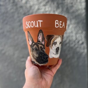 以下が含まれることがあります： 手描きで犬の肖像画が描かれたテラコッタ製の植木鉢。鉢には白いペンキで「Scout」と「Bea」の名前が書かれています。