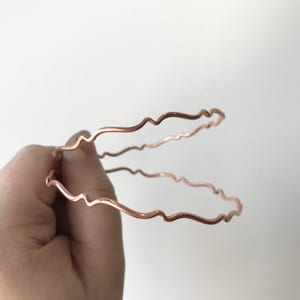 May include: Two thin, rose gold-colored metal bangles with a wavy, organic design.