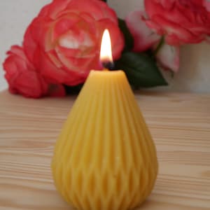 Peut inclure: Une bougie en cire d'abeille jaune en forme de poire avec une texture côtelée verticale et un motif en losange. La bougie est allumée, avec une petite flamme vive au sommet. Derrière se trouvent deux grandes roses rose et rouge vif.
