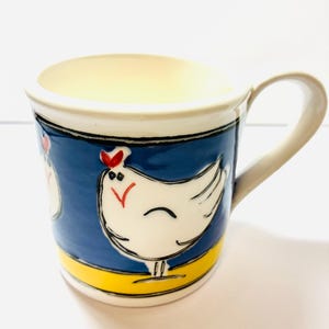 May include: A ceramic mug with a cream-coloured interior and handle. The exterior features a vibrant blue band decorated with a white chicken illustration, complete with a red comb and black outlines, set against a solid yellow base stripe.