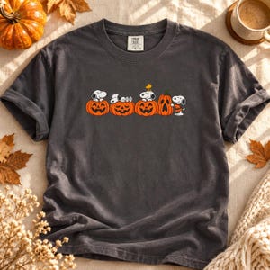 May include: A charcoal gray short-sleeve t-shirt featuring a graphic of Snoopy and Woodstock interacting with several orange jack-o'-lanterns. The shirt has a crew neckline and a tag at the inner collar that reads Comfort Colors L G G.