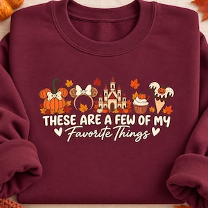 May include: A maroon crewneck sweatshirt featuring a graphic print of a pumpkin, mouse ears, a castle, a cupcake, and an ice cream cone surrounded by autumn leaves. The text reads, These are a few of my favorite things, in white and cream.