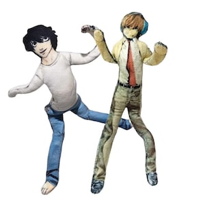 Puede incluir: Dos muñecos de tela planos e impresos que representan personajes de anime. Una figura tiene cabello negro, camisa blanca de manga larga y jeans azules. La segunda figura tiene cabello rubio, auriculares azules, camisa amarilla con corbata roja y pantalones color canela.