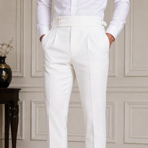 May include: White trousers featuring a high-waisted design with a wide waistband, side buckle adjusters, and double pleats at the front. These tailored pants have a straight-leg cut with cuffed hems and are paired with black leather dress shoes.