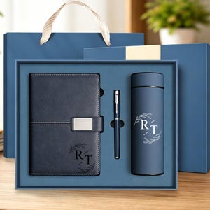 Può includere: Un set regalo blu navy contenente un taccuino con texture in pelle con chiusura in metallo argentato, una penna in metallo sottile e una borraccia termica cilindrica. Sia il taccuino che la borraccia presentano le lettere RT stampate in bianco.