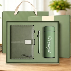 Puede incluir: Un set de regalo en color verde salvia que incluye un cuaderno con cierre plateado, un bolígrafo a juego y una botella cilíndrica aislante. El nombre Mcknight aparece impreso en letra cursiva blanca tanto en el cuaderno como en la botella.