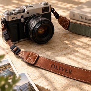 Op de afbeelding: Een zilveren en zwarte digitale camera met een bruine leren camerariem waarop de naam OLIVER in het zwart is gedrukt. De riem heeft witte stiksels, zwarte plastic verstelgespen en een groene stoffen onderkant bevestigd aan de camerabehuizing.