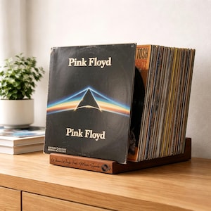 May include: A dark brown wooden vinyl record storage stand holding a collection of records. The front-most record is Pink Floyd's The Dark Side of the Moon with its iconic prism graphic. The stand features the engraved text Where Words Fail, Music Speaks.