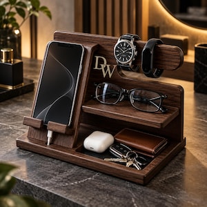 May include: A dark brown wooden desk organizer featuring a vertical smartphone stand with a charging cable slot, a horizontal bar for a wristwatch and fitness tracker, a shelf for eyeglasses, and a tray for earbuds, a wallet, and a set of keys.