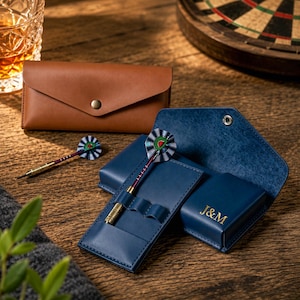 May include: A tan leather dart case with a snap closure and a navy blue leather dart case featuring the gold embossed letters J&M. The navy case holds a dart with a metallic purple shaft and a striped flight with a green and red watermelon graphic.