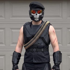 Op de afbeelding: Een zwart tactisch kostuum met een schedelmasker met oranje glazen, een zwarte baret en een mouwloos vest. De outfit bevat een patroonband met goudkleurige munitie, zwarte handschoenen, dijbeenbescherming, kniebeschermers en zwarte gevechtslaarzen.