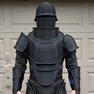May include: A full-body matte black suit of armor featuring a rounded helmet with a faceplate, a high-collared chest piece, shoulder pauldrons, forearm guards, and leg plating that covers the thighs and shins, all constructed with a smooth finish.