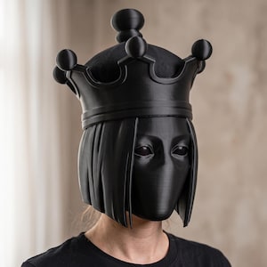 May include: A matte black full-head mask featuring a stylized bob haircut and a prominent crown topper with spherical points. The mask has smooth, solid surfaces with defined facial features, including almond-shaped eye openings and a sleek, uniform finish.
