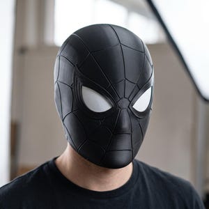 May include: A black full-head mask featuring a textured web pattern across the surface and large, almond-shaped white eye lenses. The mask covers the entire head and face, displaying a detailed grid design consistent with a superhero character.