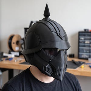 May include: A matte black full-face helmet featuring a prominent central spike on the crown, a wide visor area with a dark tinted lens, and a structured geometric faceplate with angular vents and molded panel details across the entire exterior surface.