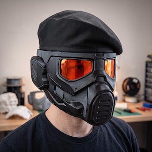 Peut inclure: Un casque tactique noir avec une finition mate, une grille de respiration frontale proéminente et de larges visières orange réfléchissantes. Le casque est surmonté d'un béret noir uni, créant une apparence sombre, structurée et cohérente.