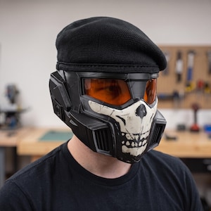 Puede incluir: Un casco táctico negro con un gráfico de calavera blanca en la placa frontal y una visera naranja translúcida. El casco está cubierto por una boina de punto negra y está construido con paneles rígidos angulares y rejillas de ventilación laterales.