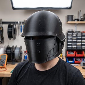May include: A matte black tactical helmet featuring a rounded dome, a wide visor opening, and a lower face guard secured with four circular rivets. The helmet has a structured, multi-piece design with visible layered textures across the surface.