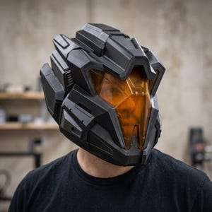 Könnte beinhalten: Ein mattschwarzer Integralhelm mit geometrischem, kantigem Design und einem markanten, durchscheinenden bernsteinfarbenen Visier. Der Helm hat ein geschichtetes, mechanisches Aussehen mit scharfen Kanten und vertieften Paneelen.