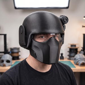 May include: A matte black tactical helmet featuring a full-face mask, a clear visor, and a side-mounted circular accessory. The helmet has a smooth, rounded dome and a structured lower section that covers the nose and mouth area completely.