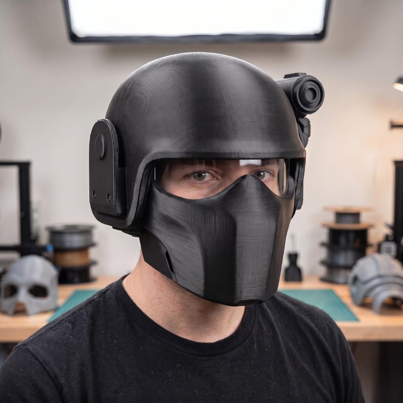 May include: A matte black tactical helmet featuring a full-face mask, a clear visor, and a side-mounted circular accessory. The helmet has a smooth, rounded dome and a structured lower section that covers the nose and mouth area completely.