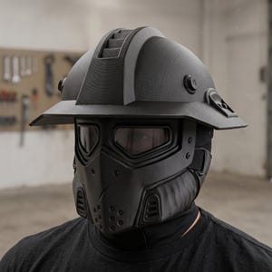 Puede incluir: Un casco táctico negro mate con ala ancha, una cresta central estriada y una máscara facial completa con protección ocular integrada. La máscara incluye una mentonera ventilada con perforaciones circulares y rejillas laterales de textura mate.