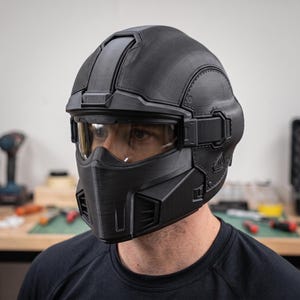 May include: A matte black full-face helmet featuring a textured, 3D-printed finish with a prominent central ridge, a wide horizontal visor with a dark tint, and a structured chin guard with integrated ventilation ports on the sides.