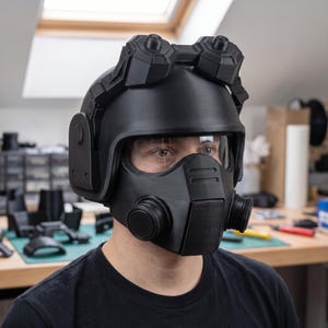 May include: A matte black tactical helmet featuring a full-face mask with a clear visor, integrated side filters, and a dual-lens goggle attachment mounted on the top. The helmet has a structured, angular design with visible 3D printed texture.