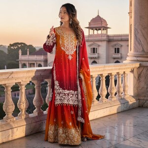 Op de afbeelding: Een set bestaande uit een tuniek met lange mouwen en een rok tot op de grond met een kleurverloop van oranje naar dieprood. Het kledingstuk is versierd met ingewikkeld zilverkleurig borduurwerk en kralen bij de halslijn en de zoom.