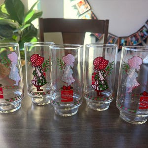 May include: A set of five clear glass tumblers featuring a graphic of a person in a bonnet holding greenery. Each glass displays the red Coca-Cola logo, a red bow, and the text Merry Christmas. The glasses have a stepped base design.