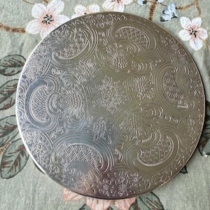 May include: A circular silver-toned metal tray featuring an intricate embossed pattern of stylized flowers, scrolling foliage, and geometric grid sections. The surface has a reflective finish with deep-set engravings that create a textured appearance.