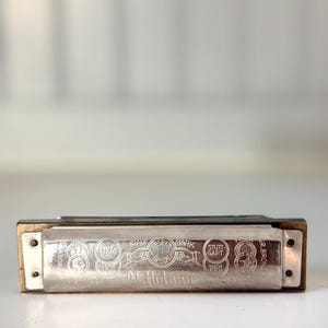 May include: A silver-toned metal harmonica with brass-colored ends. The front plate features engraved text including M. Hohner, Trade Mark, Stuttgart, 1871, and 1881, along with circular emblems depicting hands and various medals or awards.