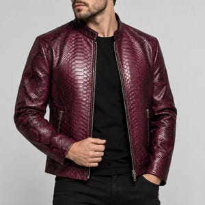 May include: A deep burgundy jacket featuring a textured snakeskin pattern, a stand-up collar, and a full-length silver-tone metal zipper closure. The jacket includes two side pockets with silver-tone zippers and long sleeves with a fitted silhouette.