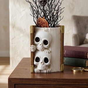 Op de afbeelding: Een decoratieve vaas in de vorm van een boek met twee witte schedels en skeletachtige handen die aan de zijkanten naar buiten komen. De vaas heeft bruine randen met goudkleurige hoekaccenten en bevat zwarte decoratieve takken en een kleine oranje glitterpompoen.