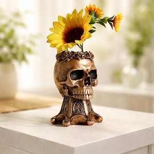 Könnte beinhalten: Eine bronzefarbene Vase in Totenkopfform mit einer Krone aus kleinen, geformten Rosen am oberen Rand. Der Sockel ist ausgestellt und mit komplexen Prägungen versehen. Drei leuchtend gelbe Sonnenblumen mit dunkelbraunen Mitten stecken darin.