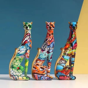 Peut inclure: Trois figurines de léopards debout arborant des motifs graffiti multicolores vibrants. Chaque sculpture présente une finition lisse et brillante avec une queue dorée distincte qui se courbe vers le haut, révélant des motifs d'art urbain.