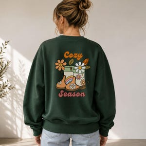Könnte beinhalten: Ein dunkelgrünes Langarm-Sweatshirt mit einem Grafikdruck auf der Rückseite. Das Motiv zeigt zwei mit Blumen und einem Regenbogen verzierte Stiefel, begleitet von dem Schriftzug Cozy Season in orangefarbenen und rosafarbenen Buchstaben.