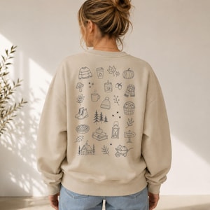 Könnte beinhalten: Ein beigefarbenes Sweatshirt mit Rundhalsausschnitt und einem schwarzen Strichzeichnungsdruck auf der Rückseite. Das Design enthält verschiedene herbstliche Symbole wie einen Pullover, einen Kaffeebecher, einen Kürbis, eine Kerze und Stiefel.