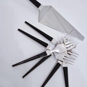 Könnte beinhalten: Ein Besteckset aus Edelstahl, bestehend aus einem Tortenheber und fünf Gabeln, jeweils mit einem eleganten schwarzen Griff. Die Utensilien sind fächerförmig angeordnet und zeigen die polierten silbernen Metallköpfe und schwarzen Griffe.