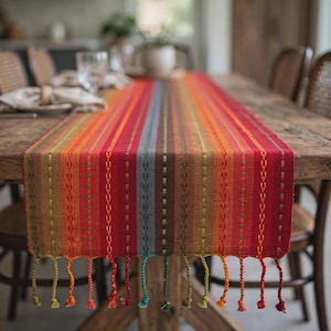 May include: A rectangular table runner featuring a vibrant vertical striped pattern in shades of red, orange, yellow, green, and blue. The fabric has a woven texture with small embroidered stitch details and finished with twisted fringe tassels.