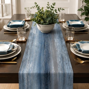 May include: A long, rectangular table runner featuring a vertical, textured gradient pattern in shades of deep navy, slate blue, and light gray. The runner is centered on a dark wood dining table set with white plates, blue napkins, and gold cutlery.