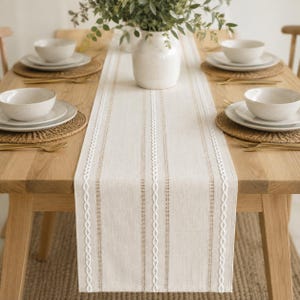 May include: A beige table runner featuring vertical white embroidered stripes and a textured fabric finish, centered on a light wood dining table with place settings including white bowls, woven placemats, and gold-toned cutlery.