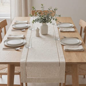 May include: A light beige linen table runner with a decorative hemstitch border, centered on a wooden dining table set with white ceramic plates, clear glass tumblers, silver cutlery, folded linen napkins, a salt shaker, and a vase of eucalyptus.