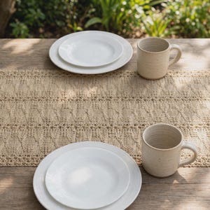 May include: A beige woven table runner with a textured lattice pattern, two white ceramic plates, and two speckled beige ceramic mugs arranged on a wooden surface. The table runner features an intricate openwork design with vertical lines.