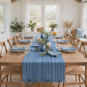 May include: A long blue woven table runner with fringe edges, placed on a wooden dining table. The runner features a textured striped pattern in various shades of blue. Matching blue cloth napkins are folded neatly at each place setting.