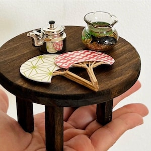 以下が含まれることがあります： 濃い茶色の木製ミニチュア丸テーブル。銀色の金属製ティーポット、黒い魚が入った小さなガラスボウル、緑、白、赤の幾何学模様が描かれた2つの装飾的な紙製扇子がテーブルの上に置かれています。