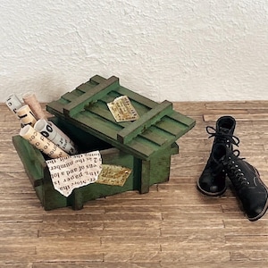 以下が含まれることがあります： 蓋が少し開いた緑色の木製ミニチュア木箱。中には小さな紙のロールがいくつか入っています。木箱の横には黒い編み上げブーツが一足置かれています。木箱の蓋と前面には小さな長方形のラベルが貼られています。