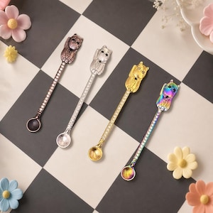 May include: Four metal spoons with long handles featuring a cat figure at the top. The handles are decorated with small clear stones. The spoons come in four distinct finishes: a dark bronze, a polished silver, a bright gold, and a rainbow iridescent.
